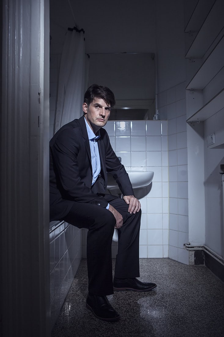 Mann in Anzug sitzt im Badezimmer vor Waschbecken, dunkle Kleidung.