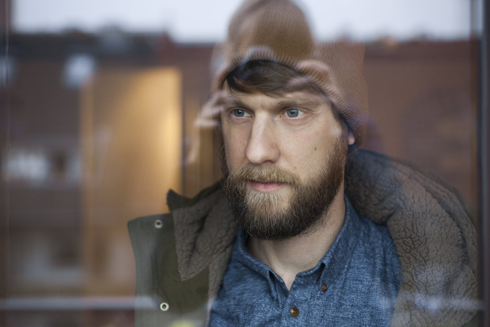 Mann mit Bart und Mütze schaut nachdenklich durch ein Fenster, urbanes Umfeld.