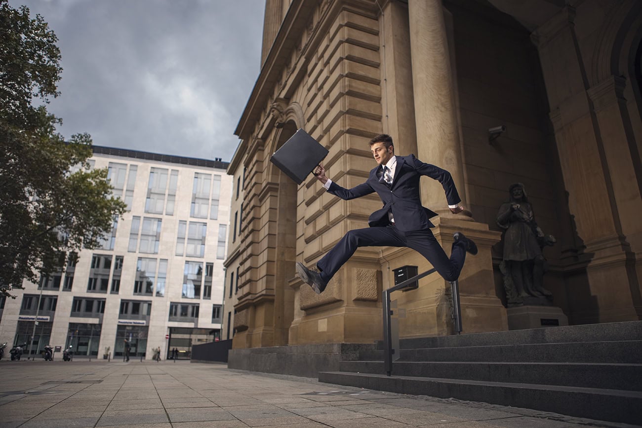 Mann im Anzug springt mit Aktentasche vor Kölner Architektur, kreative Hochzeitsfotografie.