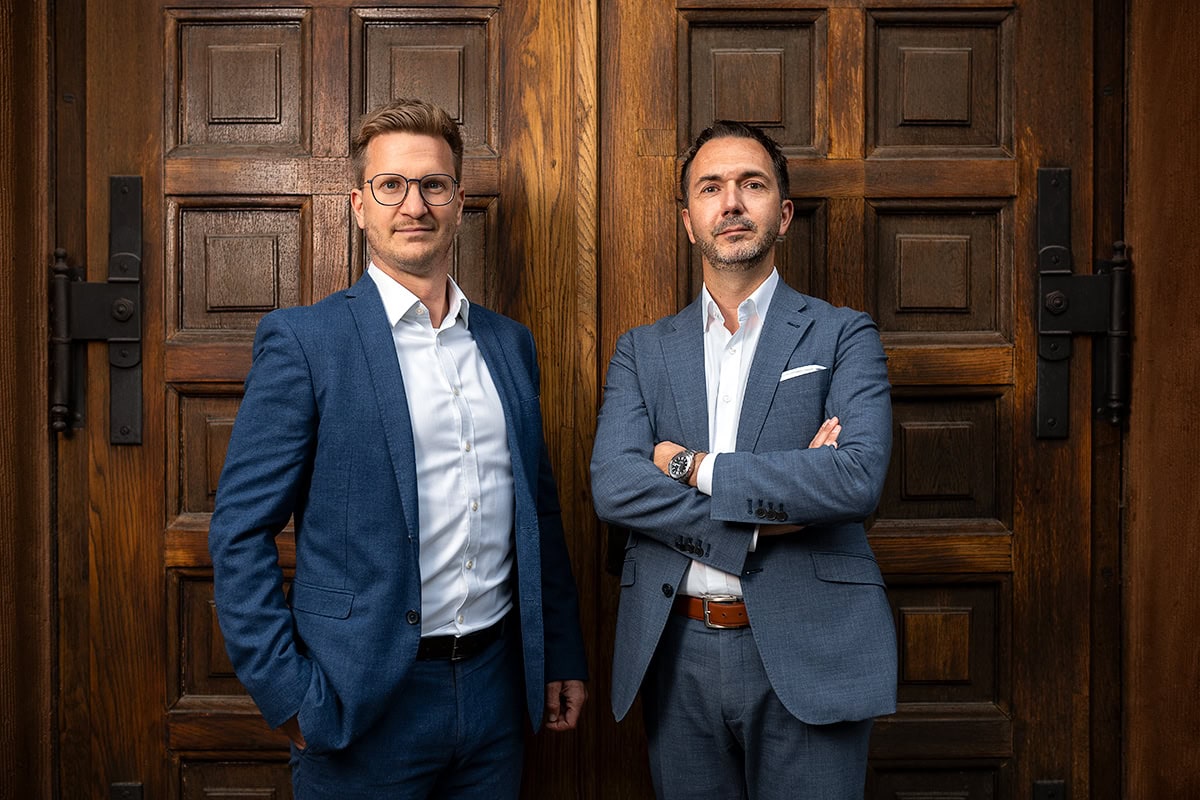 Zwei professionell gekleidete Männer in Anzügen vor einer Holzvertäfelung.