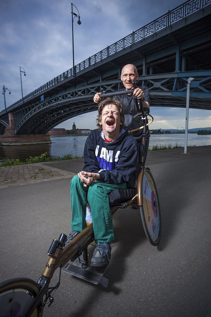 Zwei lachende Männer auf einem Fahrrad unter der Hohenzollernbrücke in Köln.