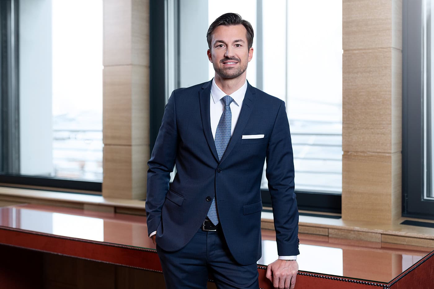 Geschäftsmann im Anzug vor Fenster mit Blick auf die Stadt, professionelle Business-Fotografie.