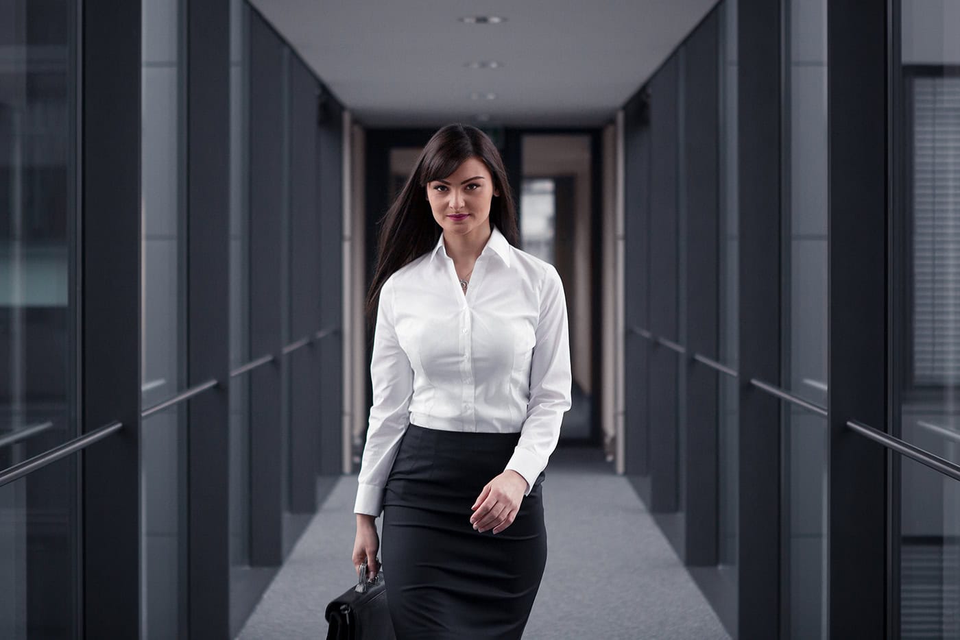 Junge Businessfrau in weißer Bluse und Bleistiftrock geht selbstbewusst durch einen modernen Büroflur