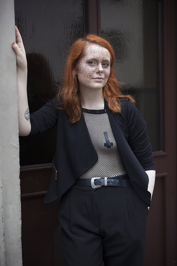 Eine junge Frau mit leuchtend roten Haaren, Sommersprossen und einem Tattoo am linken Unterarm steht lächelnd und selbstbewusst vor einer dunklen Tür. Sie trägt ein schwarzes Sakko über einem Netz-Top, eine schwarze Hose und einen Gürtel mit großer Schnalle.