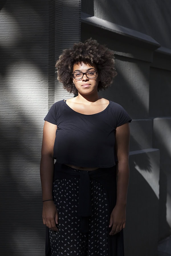 Eine junge Frau mit Afro-Frisur und Brille steht ernst und nachdenklich vor einer strukturierten grauen Wand, die teilweise von einem starken Schattenwurf bedeckt ist. Sie trägt ein schwarzes T-Shirt und ein gemustertes Kleid.