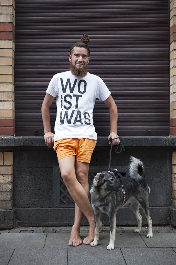 Ein junger Mann mit langem Bart, einem Zopf auf dem Kopf und orangefarbenen Shorts steht barfuß an einer braunen Rolladenwand. Er trägt ein weißes T-Shirt mit dem Aufdruck "WO IST WAS" und hält die Leine eines grauen Hundes, der neben ihm sitzt.