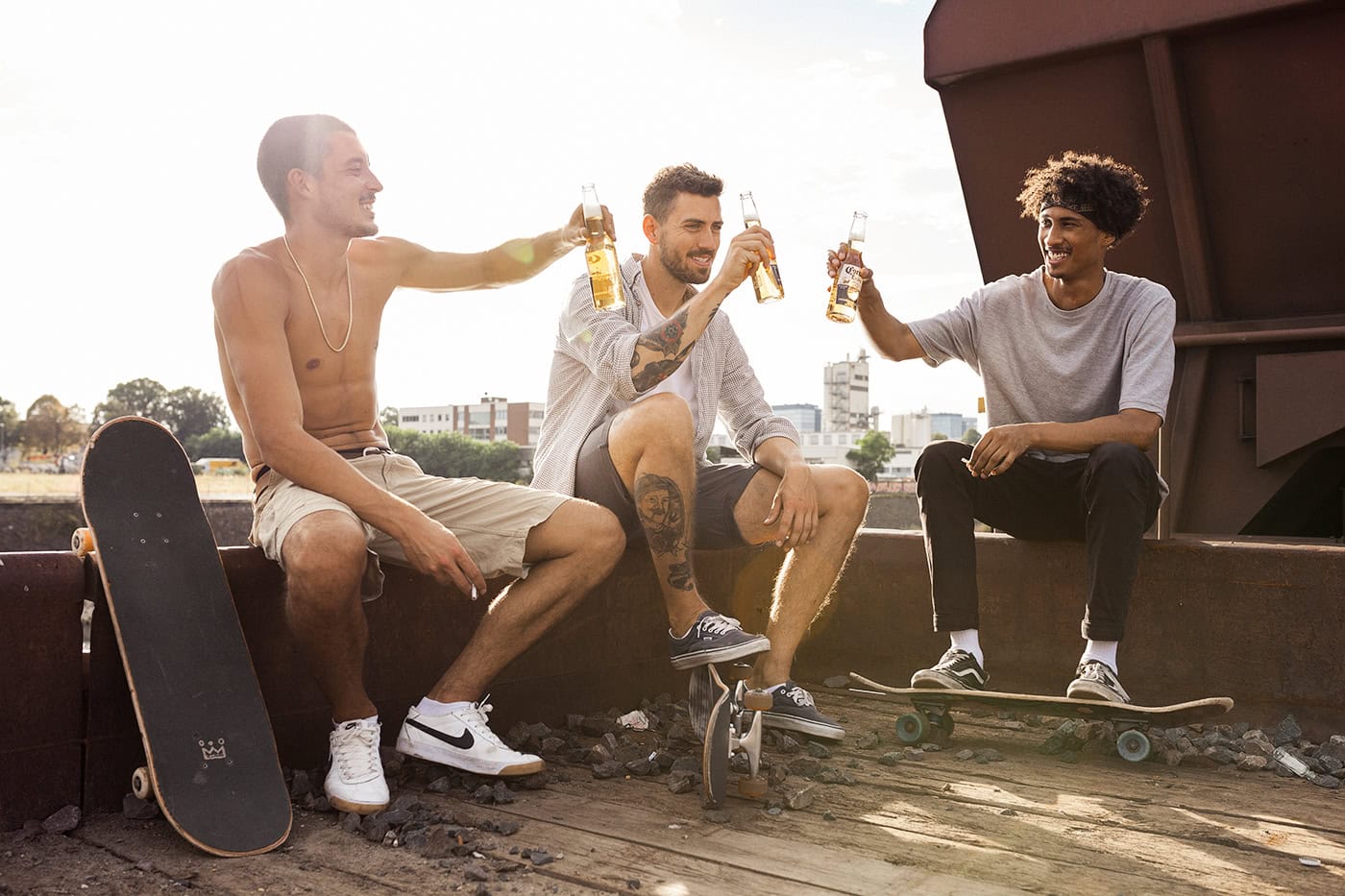 Junge Männer trinken Bier und feiern auf einem Dach mit Skateboards, Sonnenuntergang im Hintergrund.