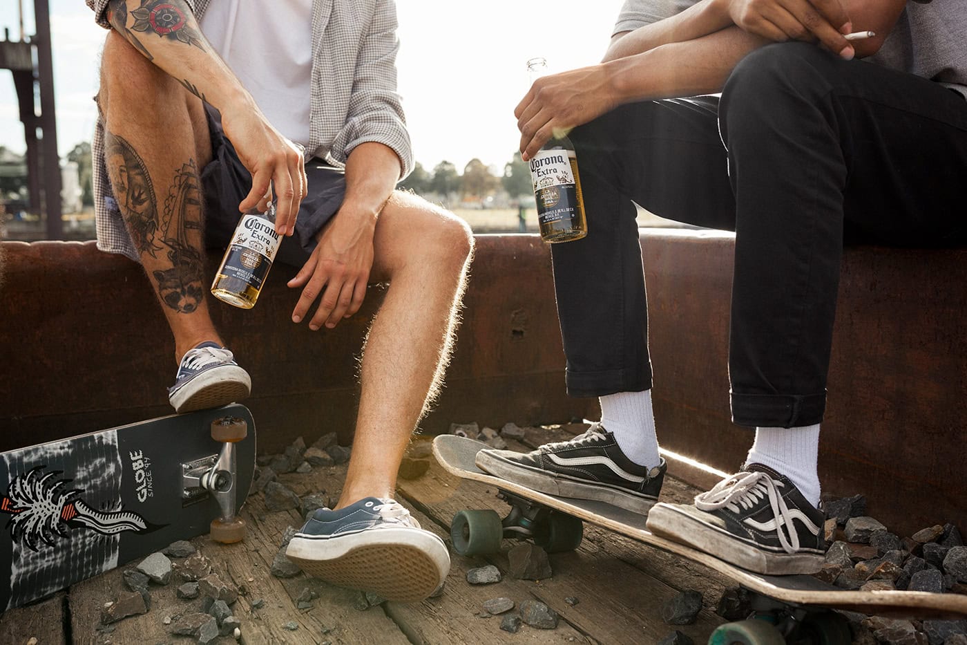 Zwei Freunde mit Skateboards und Getränken sitzen am Bahnübergang, entspannte Atmosphäre.