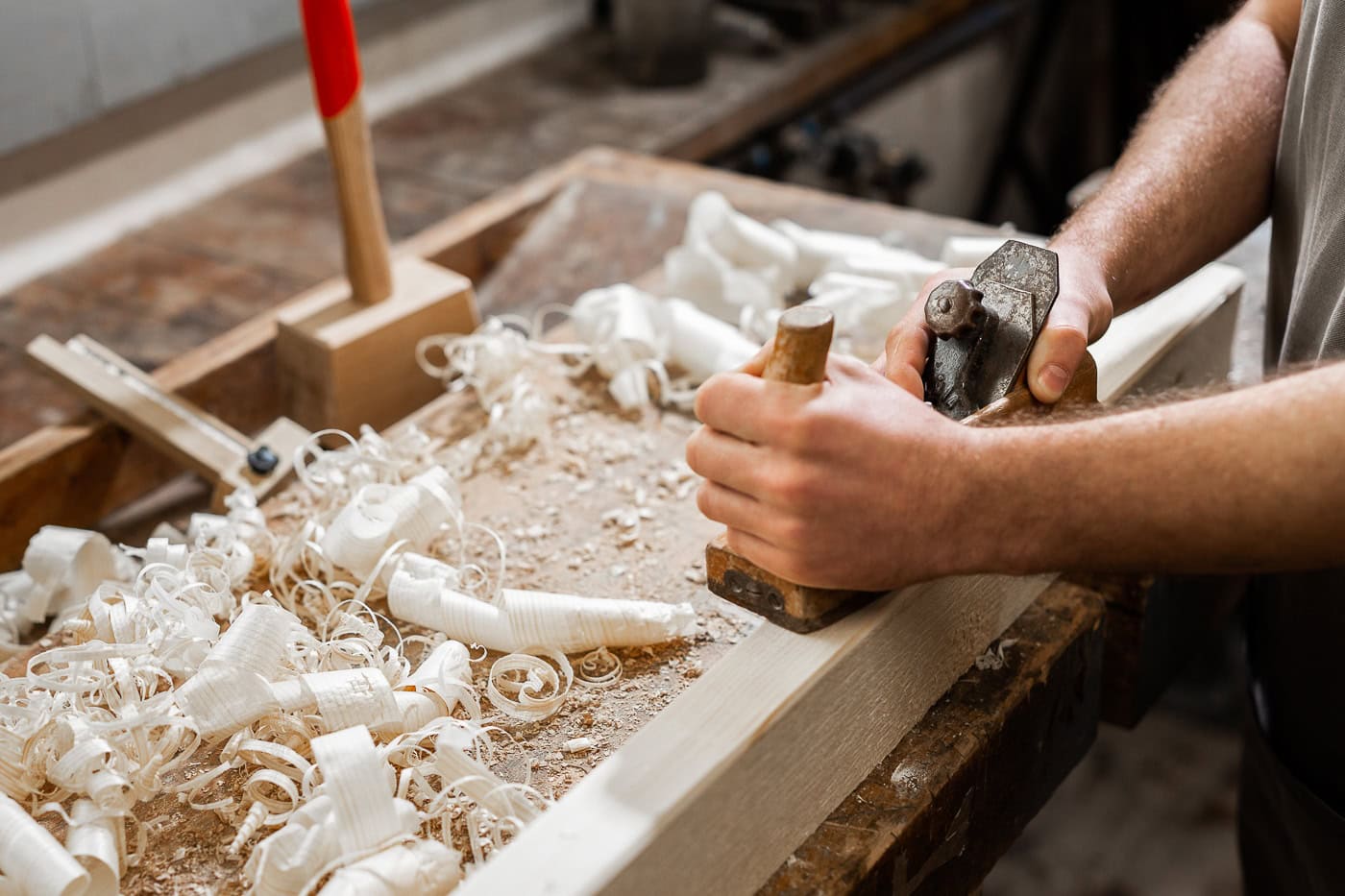 Handwerker bearbeitet Holz mit Hobel in Werkstatt, Holzspäne sichtbar.