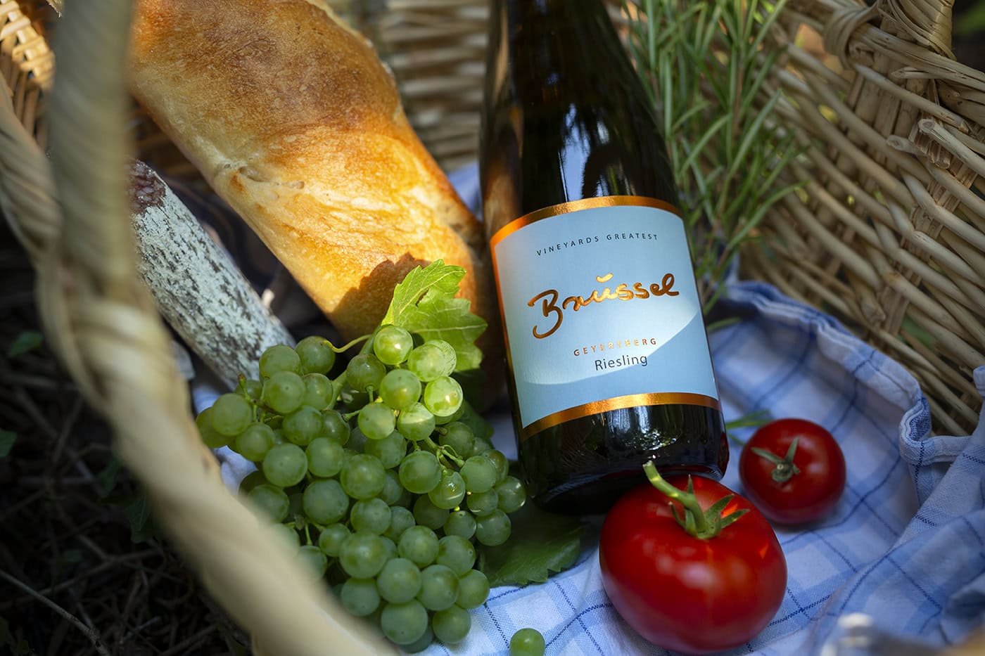 Ein rustikaler Weidenkorb ist mit einer Flasche "Brüssel" Riesling, einem Baguette, grünen Weintrauben und zwei roten Tomaten gefüllt. Ein kariertes Tuch liegt darunter. Das Ganze ist draußen im Sonnenlicht platziert, möglicherweise in einem Weinberg, da grüne Blätter und Ranken angedeutet sind.
