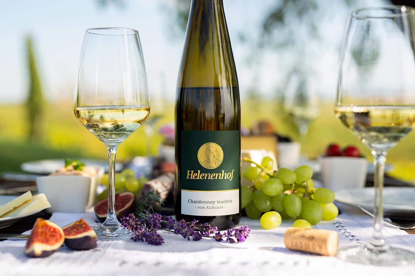 Eine grüne Weinflasche mit dem Etikett "Helenenhof Chardonnay trocken" steht auf einem Tisch im Freien. Daneben befinden sich zwei gefüllte Weingläser, eine Schale mit grünen Trauben, geschnittene Feigen, Lavendelzweige und ein Korken. Im Hintergrund sind undeutlich Rebstöcke zu erkennen.