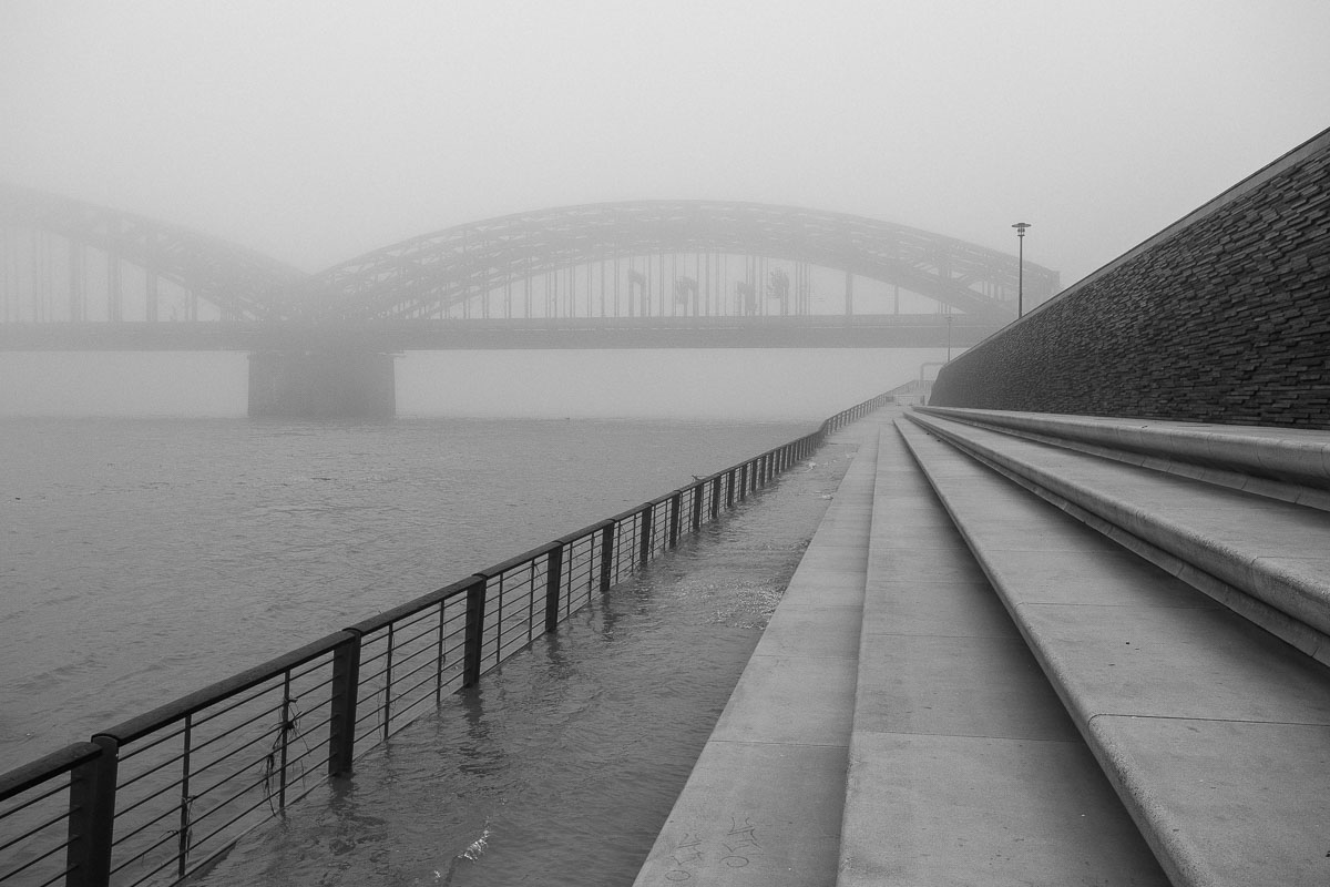 Nebelverhangene Rheinpromenade in Köln mit Brücke im Hintergrund.