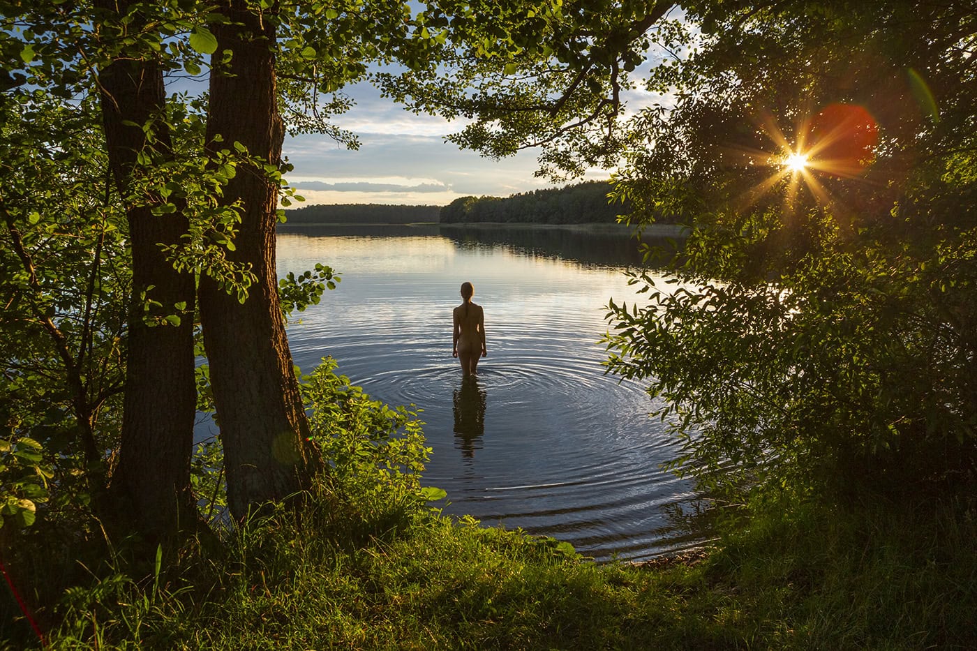 Frau steht im Wasser eines Sees bei Sonnenuntergang, umgeben von Bäumen.