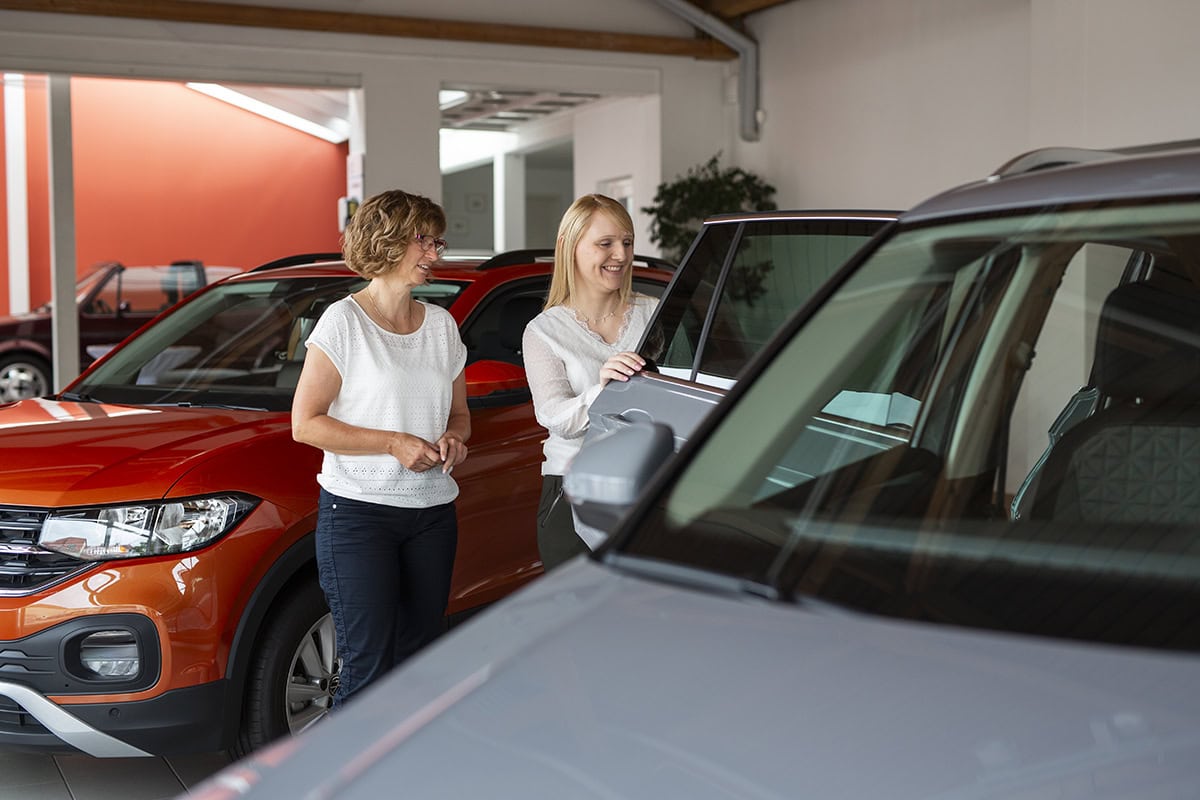 Professionelle Autoberatung: Kundin und Verkäuferin im Gespräch am Neuwagen. Im Hintergrund sind weitere helle Autos und der helle Showroom eines Autohauses zu sehen.