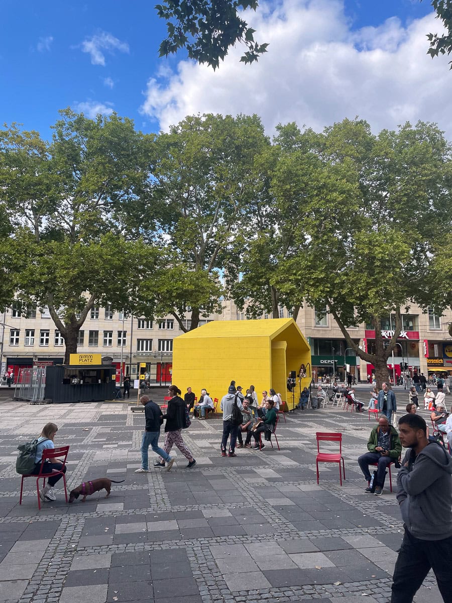 Straßenansicht in Köln mit gelber Kunstinstallation, Bäumen und Passanten.